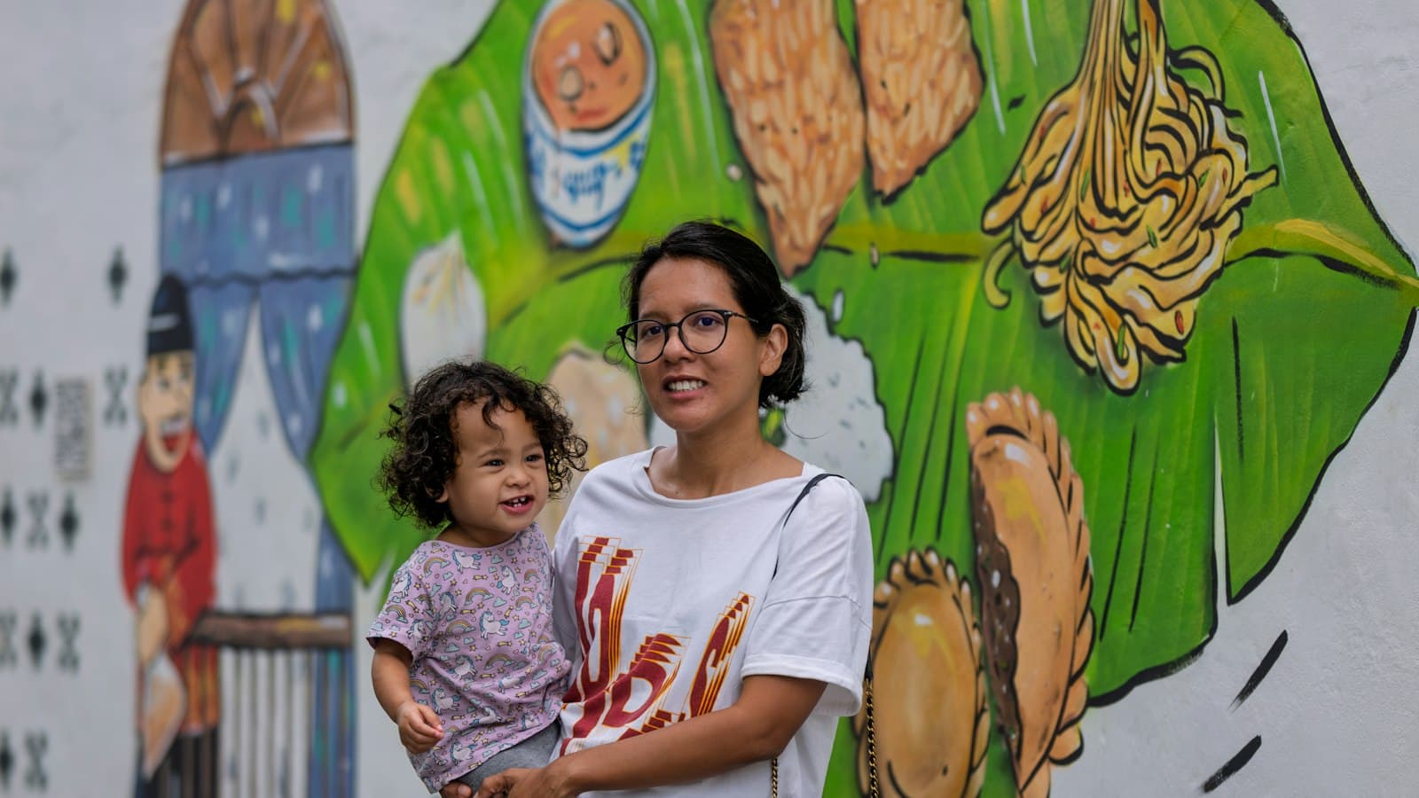 A mother holding her young daughter in front of a food mural
