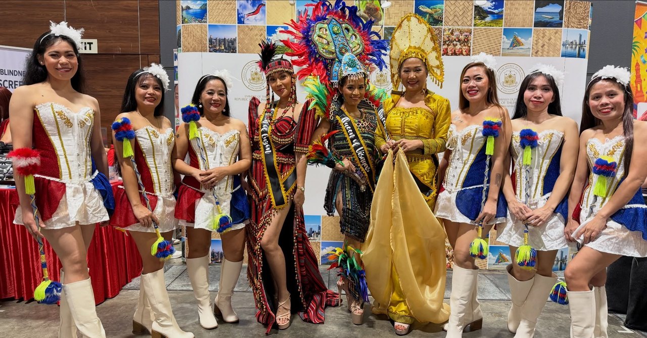 Filipino cultural performers at Fiestang Pinoy 2026, including majorettes in red-and-blue and white uniforms alongside pageant queens in elaborate festival costumes representing Luzon and the Visayas, in front of a Philippine tourism backdrop